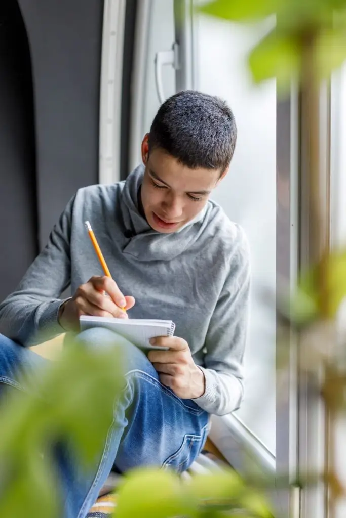 Portrait of diligent teenage boy sitting on the window sill, writing homework and studying for school.