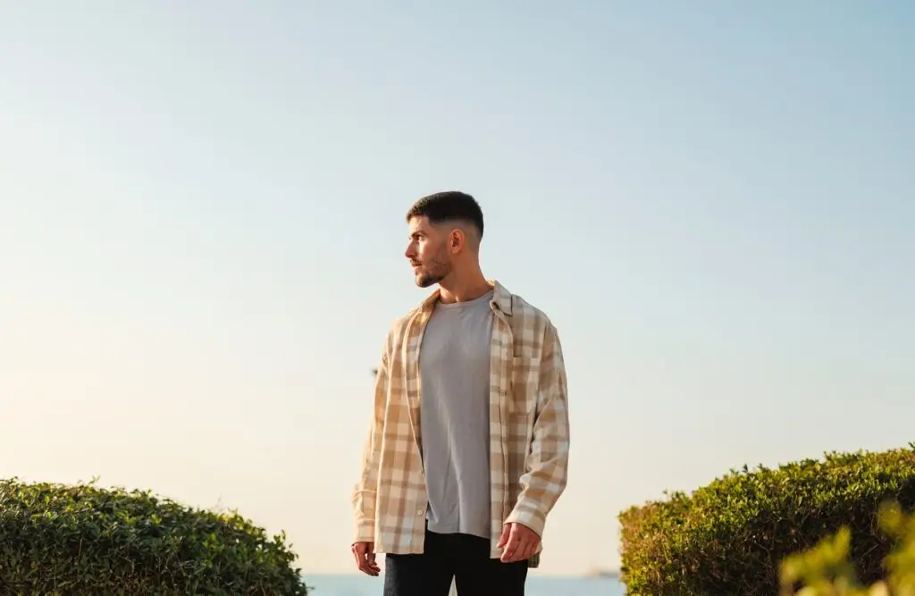 A young Caucasian man stands outdoors, looking to the side with a serene sky in the background. Dressed in casual clothing, he exudes happiness and a carefree lifestyle, embodying vitality and enjoyment of nature.